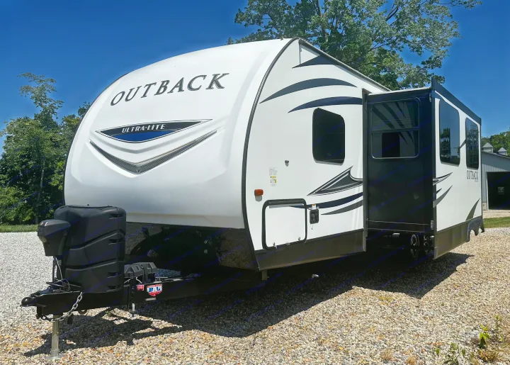 A white Outback Ultra-Lite travel trailer, suitable for RV rental, is parked on gravel under a blue sky with its door open.. Keystone RV Outback Ultra-Lite 2018