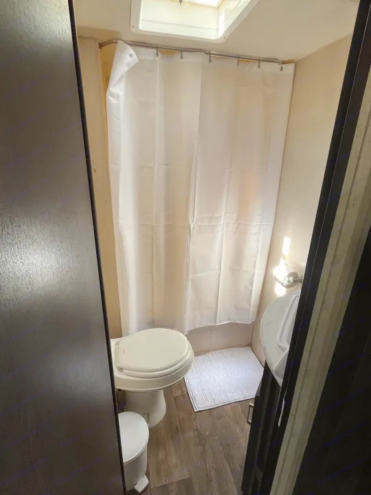 An RV rental bathroom: compact design with toilet, shower curtain, and bath mat on wood-look flooring.. Forest River Wildwood X-Lite 2018