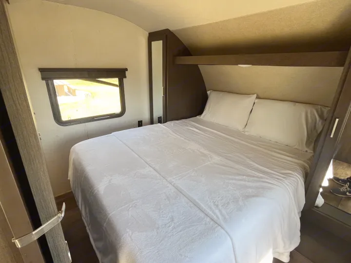 Cozy rv rental bedroom features a queen bed with white linens, a bright window view, and built-in cabinets.. Forest River Wildwood X-Lite 2018