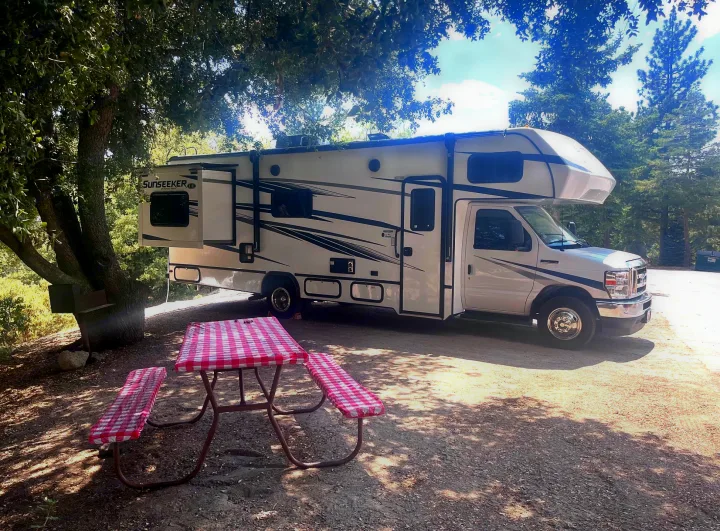 White RV rental at a shaded campsite with a red checkered picnic table and BBQ. Perfect for outdoor camping adventures!. Forest River Sunseeker LE 2023