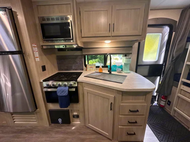 RV rental kitchen featuring light wood cabinets, stainless steel fridge, microwave, gas stove, and sink on light countertops.. Forest River Aurora 28BHS 2024