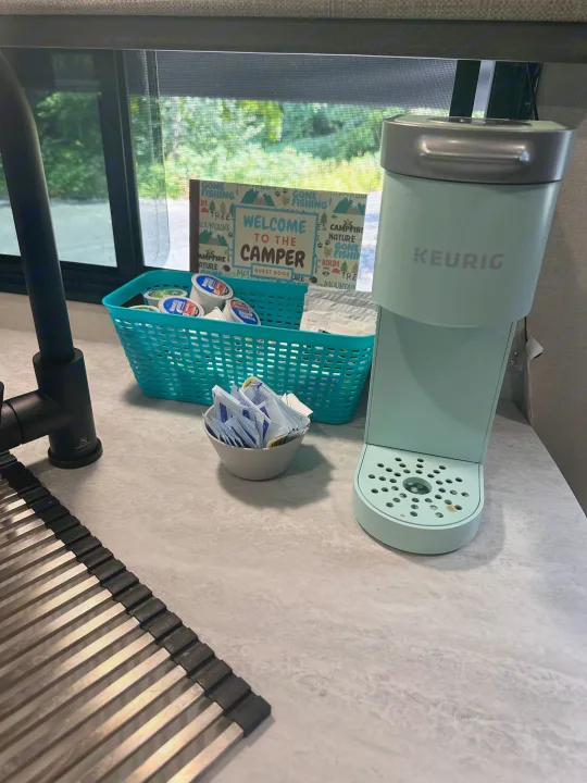 An RV rental offers a handy coffee station: a mint green Keurig, coffee pods, and sweeteners, with a camper welcome sign.. Forest River Aurora 28BHS 2024
