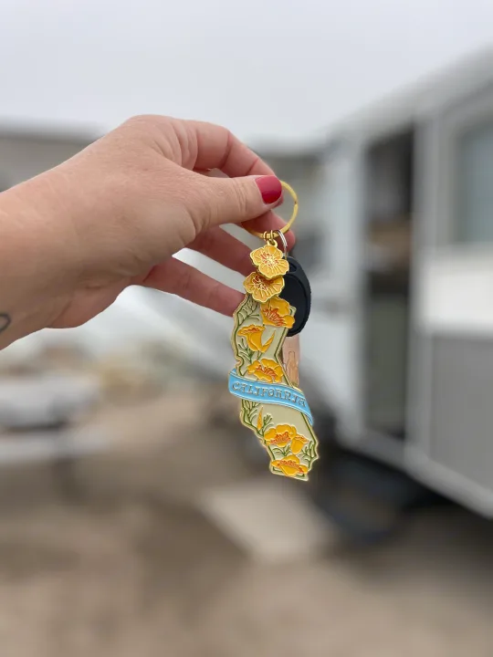 A hand holds a California poppy keychain and RV keys, ready for an RV rental adventure. An RV is visible in the background.. Forest River Wildwood 2011