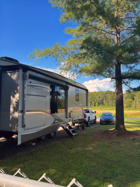 Spacious RV rental parked with truck & car under a blue sky, beside a large pine tree, field, & forest.. Jayco Eagle 2020