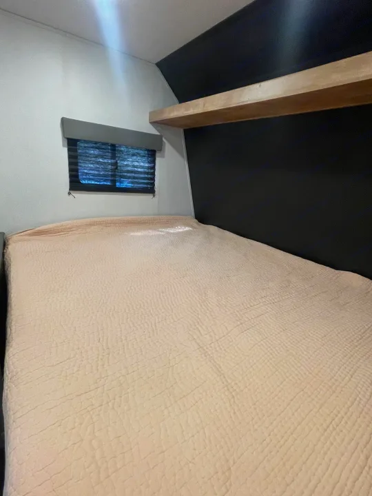RV rental bedroom featuring a cozy, textured bed, window with blinds, and modern black wall with a wooden shelf.. Keystone RV Springdale 2022