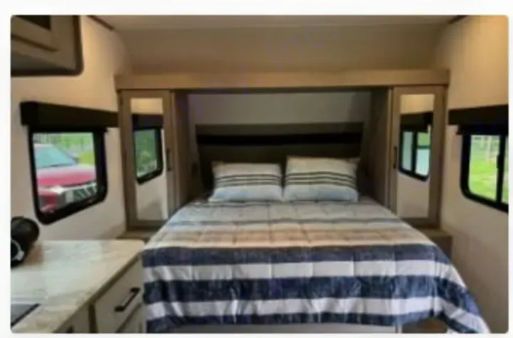 Inside an RV rental bedroom: A comfortable queen bed with striped bedding, mirrored storage, and windows. Perfect for travel!. Dutchmen Kodiak SE 2024