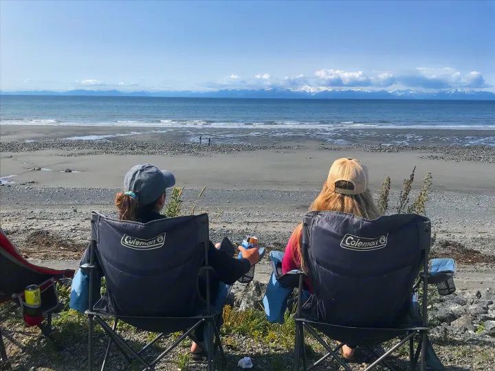 Two people in camping chairs gaze at a vast beach, ocean, and distant snowy mountains. Perfect relaxation after an RV rental journey.. Thor Four Winds 2019