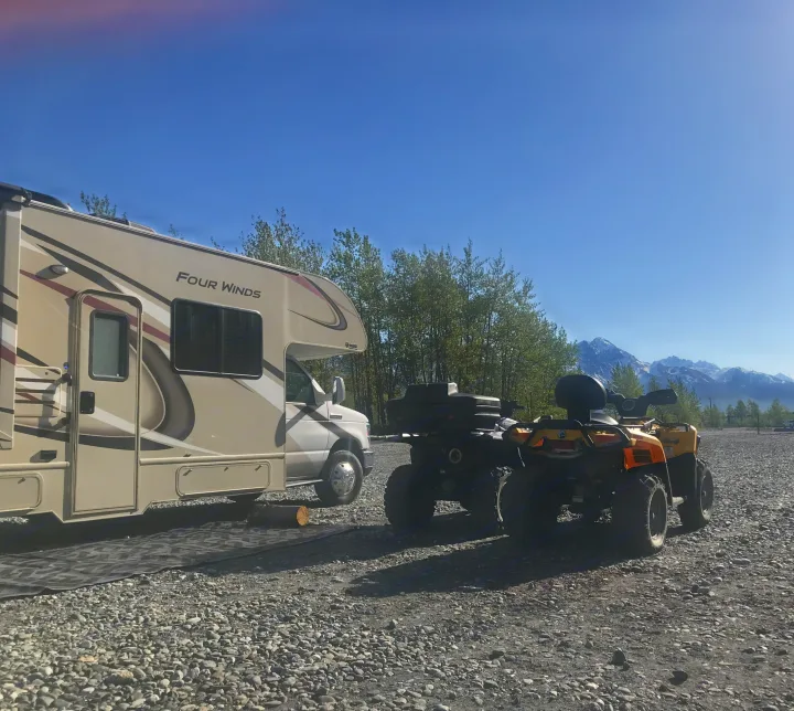 RV rental and ATVs parked on a gravel campsite with stunning mountains in the background.. Thor Four Winds 2019