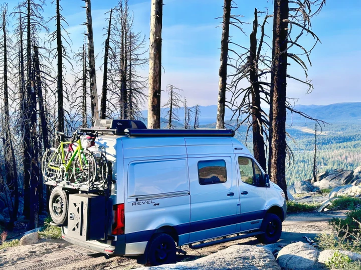 Adventure-ready RV rental, loaded with bikes, parked among stark, tall trees, overlooking a vast, mountainous landscape.. Winnebago Revel 2023