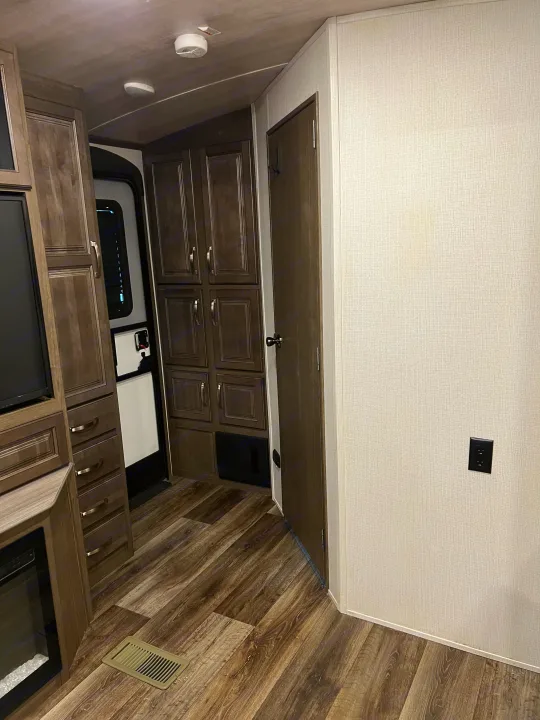 An RV rental interior showing dark wood-look cabinetry, a flat-screen TV, durable flooring, and a light-colored wall with an outlet.. Keystone RV Cougar 2023