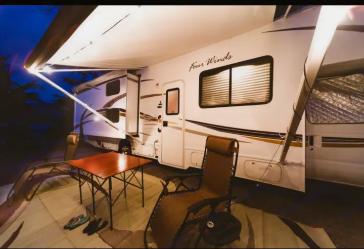 Cozy RV rental campsite at night, featuring a brightly lit awning, outdoor table, and comfy chairs on a rug.. Other Four Winds 2013