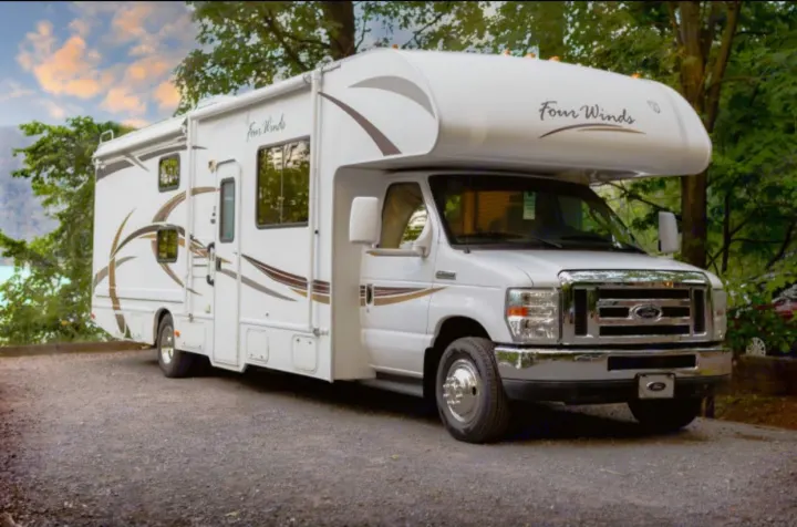 A white Four Winds Class C RV rental, with an extended slide-out, parked on gravel amidst trees. Ideal for road trips & camping.. Other Four Winds 2013