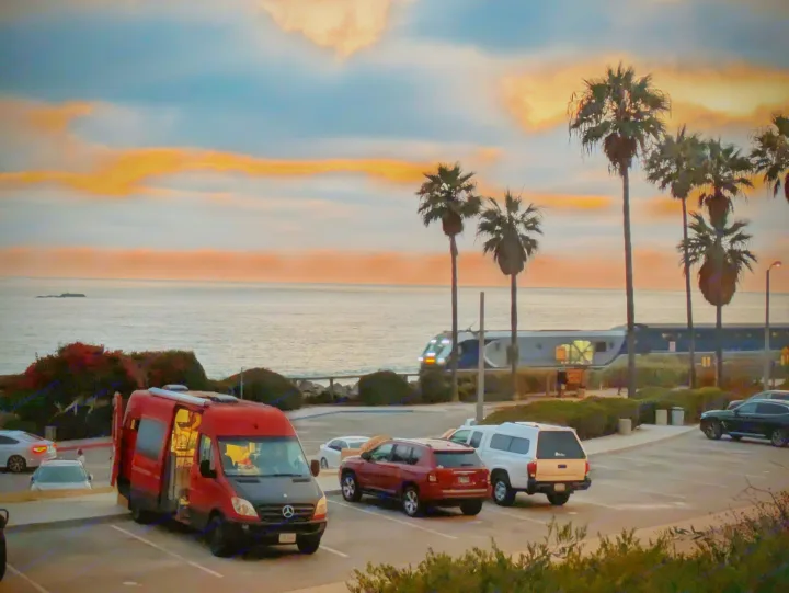 Red RV rental by the ocean at sunset, with palm trees and a train passing. A beautiful coastal scene.. Mercedes-Benz Sprinter 2008