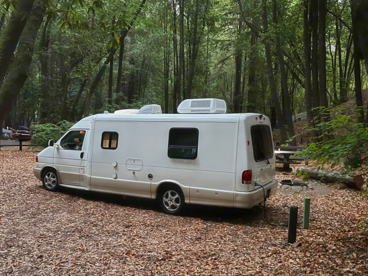 This white rv rental is nestled in a shady, leaf-covered forest campground, featuring a picnic area and utility hookups.. Winnebago Rialta 2003