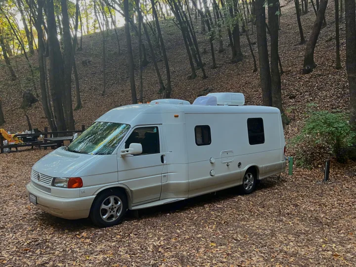 White rv rental camper van parked on autumn leaves in a peaceful, sunlit forest campsite.. Winnebago Rialta 2003