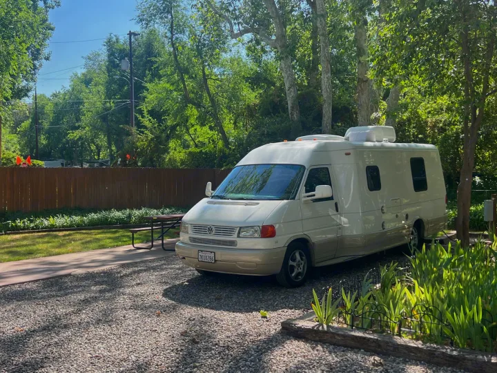A cream Class B RV parked on a gravel campsite next to a picnic table, perfect for your next rv rental.. Winnebago Rialta 2003
