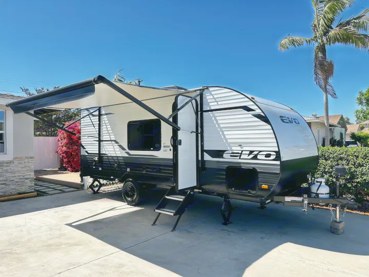 A modern white & black EVO travel trailer, with its awning extended, parked on a sunny driveway, ready for RV rental.. Forest River EVO 2024
