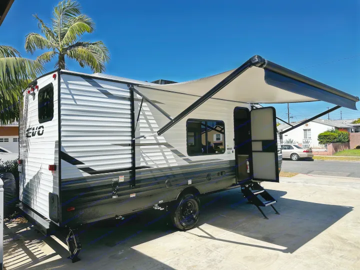 Modern white travel trailer available for RV rental, parked with awning out & door open under clear sky.. Forest River EVO 2024