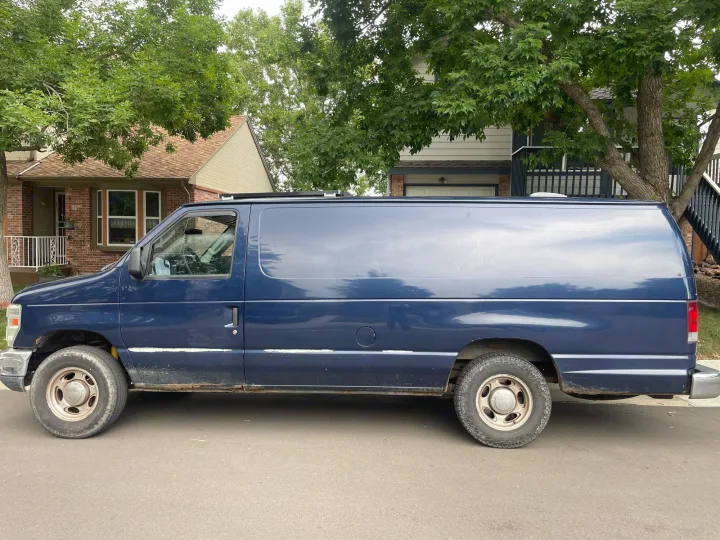 Blue van, a potential RV rental, parked on a residential street. Features a heart drawn in dust and rust along the bottom.. Ford Econoline 2010