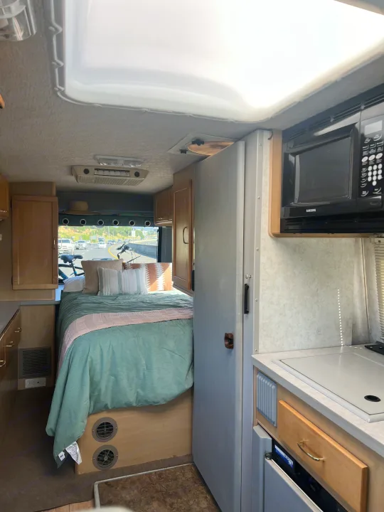 Cozy RV rental interior featuring a teal-covered bed and compact kitchen space, ideal for road trip adventures.. Winnebago Rialta 2003