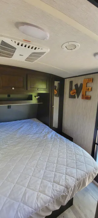 Cozy RV rental bedroom with a quilted white bed, dark wood overhead cabinets, and an AC unit, featuring "LOVE" decor.. Heartland Sundance 2019