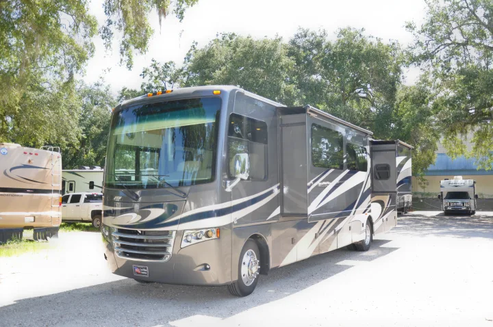 Front view of a large, modern grey rv rental motorhome, with its side slide-out extended, parked on a gravel lot under trees, with other RVs nearby.. Thor Motor Coach Miramar 2017