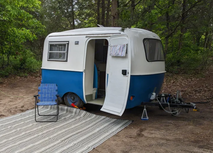 Vintage blue and white travel trailer RV rental at a wooded campsite, featuring an open door, outdoor mat, and folding chair.. Boler Boler 1300 1974