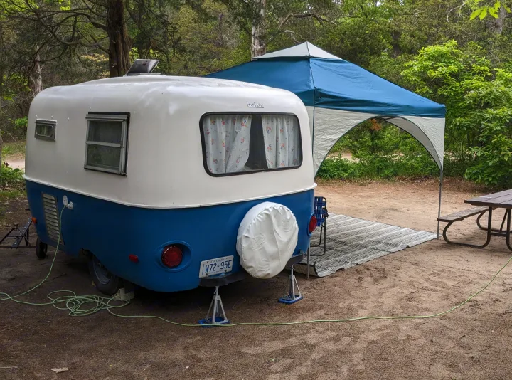 Compact blue & white RV rental set up at a shady campsite with a canopy, outdoor rug, and picnic table.. Boler Boler 1300 1974