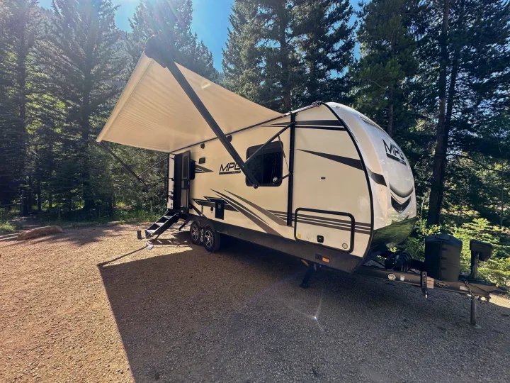 RV rental travel trailer with extended awning parked at a sunny campsite amidst tall pine trees. Perfect for outdoor adventures.. Cruiser RV MPG Ultra Lite 2022
