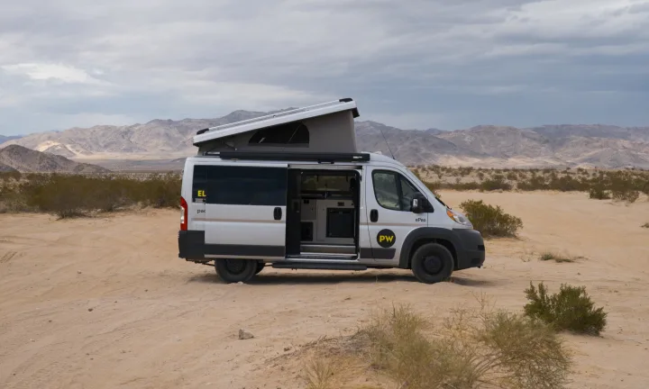 This electric RV rental camper van, pop-top open, is parked in a sandy desert with distant mountains under a cloudy sky.. Ram Pleasure Way 2019
