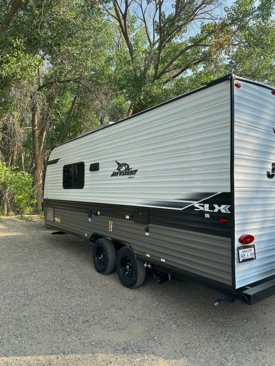 A sleek, white and grey Jayco Jay Flight SLX travel trailer, available for RV rental, is parked on a gravel lot under trees.. Jayco Jay Flight SLX 2024