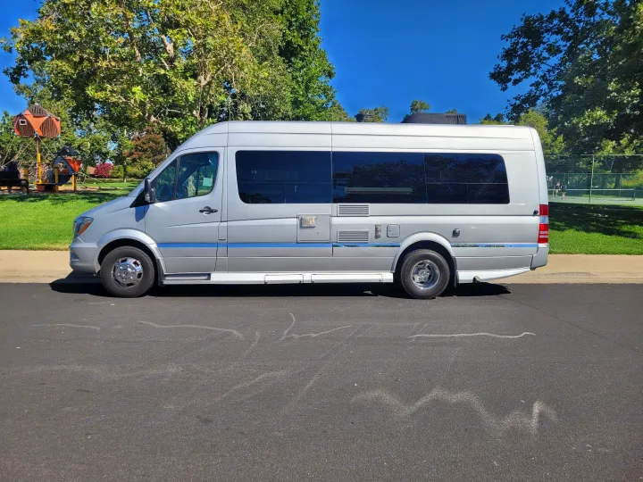 Silver RV rental parked on a sunny day. A park with a playground and tennis courts is visible nearby.. Winnebago Era 2016