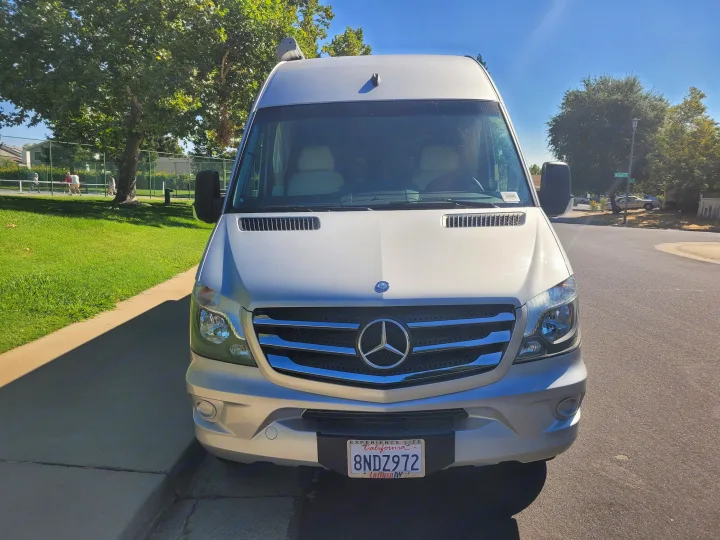 Front view of a silver Mercedes-Benz Sprinter RV rental van, parked on a sunny day with a park and residential street in background.. Winnebago Era 2016