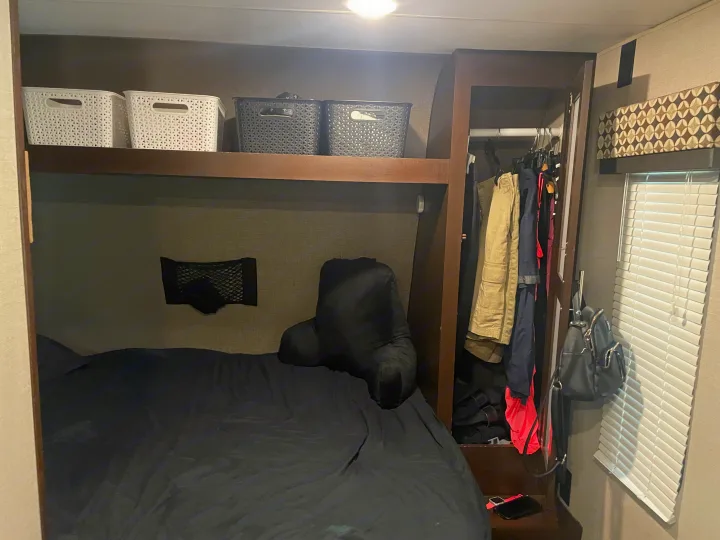 RV rental bedroom interior: dark bed, shelf storage with baskets, closet with clothes, and window blinds.. KZ Sportsmen 2019