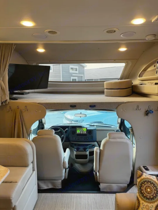 Inside an **rv rental**: driver's area, overhead bunk bed with TV, and main living space. Brightly lit and ready for travel.. Jayco Greyhawk 2021