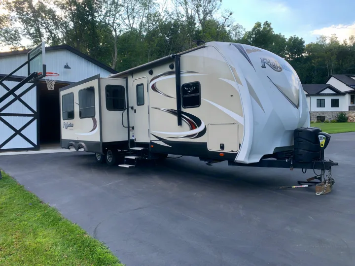 Extended slide-outs on this fifth-wheel RV, an ideal rv rental, parked on a paved driveway next to a barn and house.. Reflection BHT33 2018