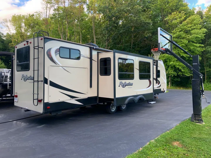 Large beige Reflection RV rental with extended slide-outs, parked on a paved driveway near a basketball hoop and trees.. Reflection BHT33 2018