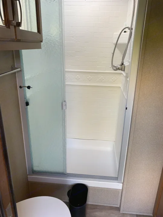 Inside an RV rental: a clean, compact bathroom with a white shower stall & frosted glass door. Toilet visible left.. Reflection BHT33 2018