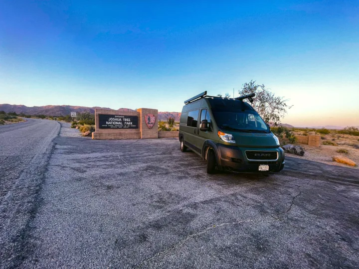 This green RV rental is parked at the Joshua Tree National Park entrance sign under a clear desert sky.. Ram Promaster 2020