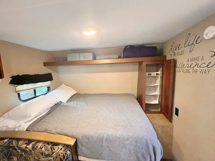 Cozy RV rental bedroom featuring a bed with gray blanket, shelving, small window, and inspirational wall art.. Keystone RV Hideout 2017