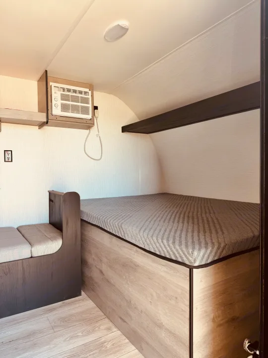 This RV rental interior features a cozy bed with a patterned mattress, a wall-mounted AC unit, and compact seating.. Cruiser RV Avenir 2024