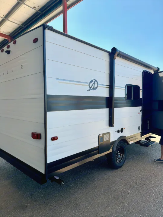 A white Avenir travel trailer, ideal for RV rental, with black trim and an awning, is viewed from its rear-right side.. Cruiser RV Avenir 2024