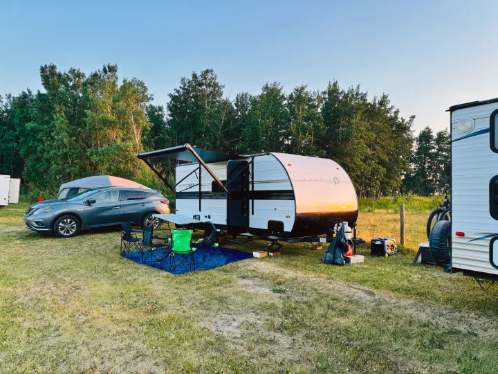 RV rental camper setup with a gray SUV, tent, chairs, and generator at a sunny, tree-lined campsite.. Cruiser RV Avenir 2024