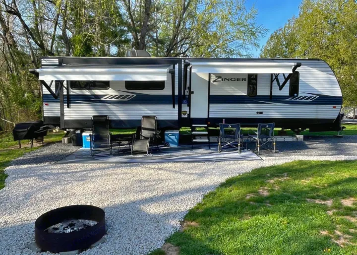 Spacious Zinger RV rental setup at a scenic campsite. Features an awning, chairs, grill, and fire pit on gravel, perfect for outdoor adventures.. Crossroads RV Zinger 2020