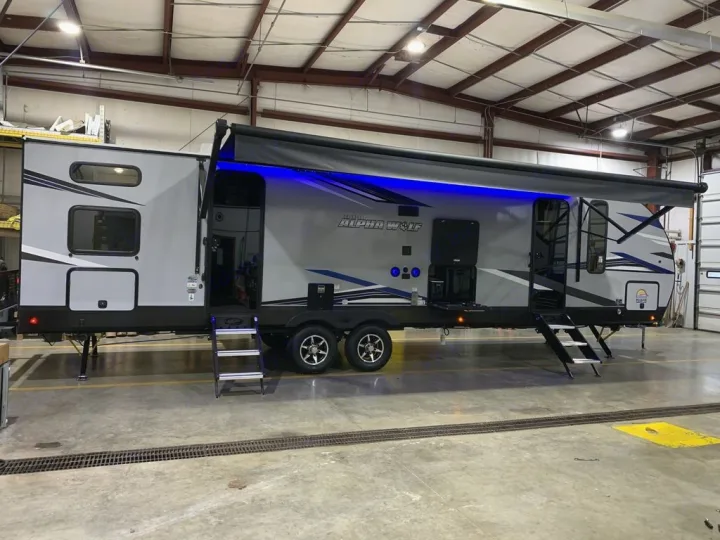This spacious grey Alpha Wolf RV, with blue-lit awning and extended steps for easy entry, is shown indoors. Consider it for your next rv rental.. Forest River Alpha Wolf 2022