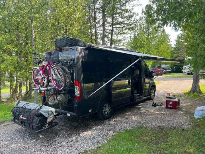 RV rental ready for a cozy stay: dark camper van with bikes, cargo, and extended awning at a gravel campsite.. Thor Tellaro 2024