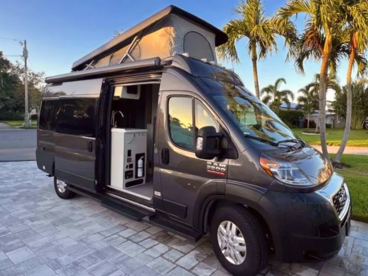 Dark grey pop-top RV rental van on a paved driveway with its side door open, revealing a compact, functional interior.. Thor Tellaro 2024