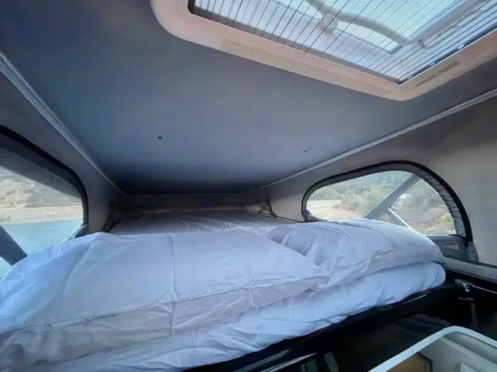 Experience comfort in this pop-top RV rental! A bed with white linens, large windows, and skylight offer scenic views.. Thor Tellaro 2024