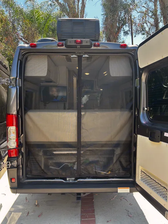 Rear view of an RV rental with open doors and a mesh screen, revealing a cozy interior with a bed and TV.. Thor Tellaro 2024
