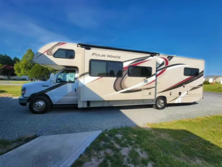 A beige Four Winds Class C motorhome, ideal for an rv rental, parked on gravel under a clear sky.. Thor Four Winds 2020